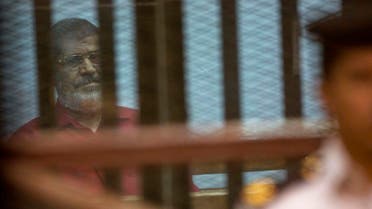 Ousted Egyptian President Mohammed Morsi, wearing a red jumpsuit that designates he has been sentenced to death, is guarded by a policeman as he sits inside a defendants cage in a makeshift courtroom at the national police academy, in an eastern suburb of Cairo, Saturday, April 23, 2016. AP
