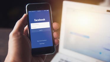 Facebook App on iPhone with computer laptop background closeup male hand hold social network on smart device concept. - Stock image...