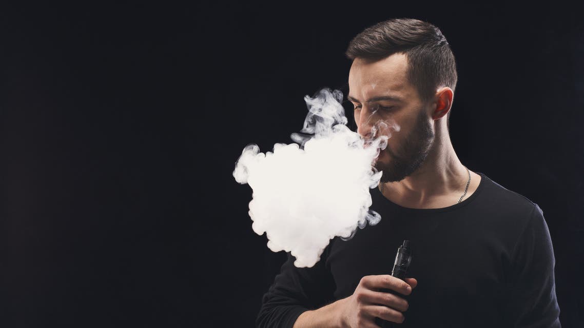 Young man vaping e-cigarette with smoke on black - Stock image