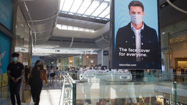 Shoppers wearing protective face masks walk past an advertising screen promoting face coverings in Westfield shopping centre in Stratford, east London on June 22, 2020. Britain's current social distancing guidelines set the distance between each person at two metres to avoid the risk of contamination to coronavirus. There is pressure on the government to reduce this distance in order to give a boost to bars, restaurants and hotels, which are scheduled to reopen next month. 