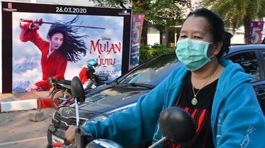 A woman, wearing a facemask amid concerns about the spread of the COVID-19 novel coronavirus, rides a motorcycle past a poster for the Disney film Mulan in Vientiane on March 11, 2020. (Mladen Antonov/AFP)