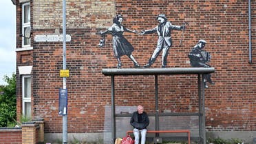 A man waits at a bus stop below a graffiti artwork of a couple dancing to an accordion player, which bears the hallmarks of street artist Banksy, on a wall in Great Yarmouth on the East coast of England on August 8, 2021. Banksy, Britain's most famous street artist, on August 13 confirmed what many had already suspected -- that he is indeed the author of several works that have recently appeared in British seaside towns. An Instagram video clip just over three minutes long, and titled A Great British Spraycation, shows the elusive artist taking a summer road trip in a beat-up camper van and with cans of spray paint stashed in a cooler.
